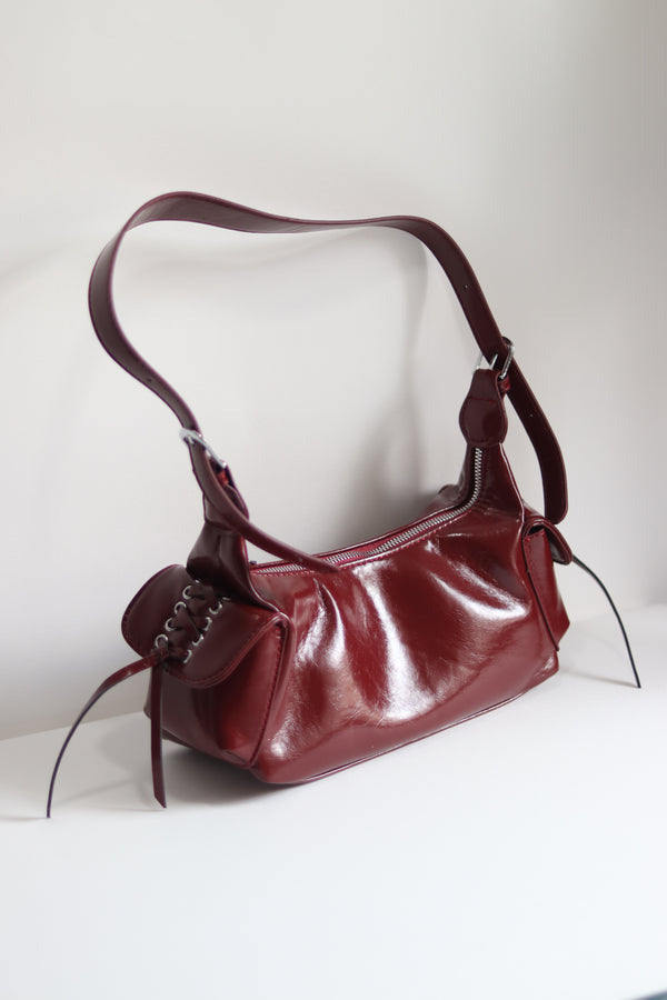 Red glossy hobo shoulder bag with adjustable strap, side lace-up pockets, and sleek zip closure design